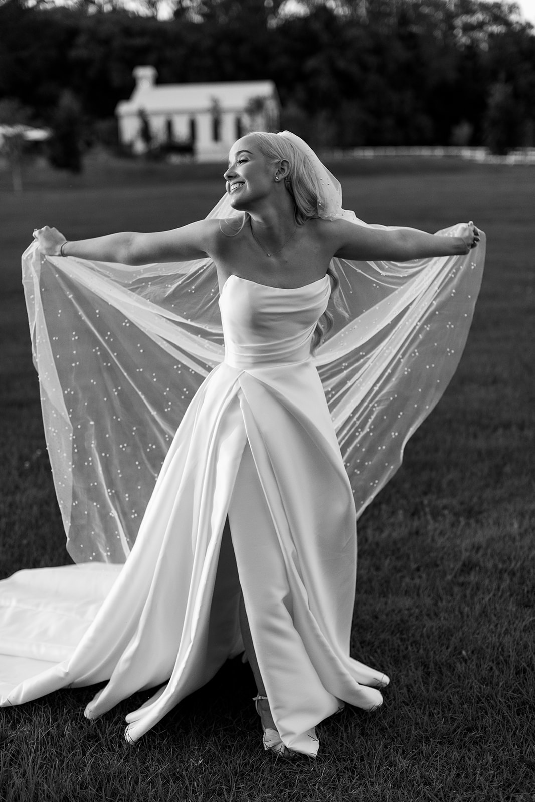 Elegant bride smiling and holding her pearl-detailed veil in the gardens of The Valley Estate, wearing a modern satin gown for her luxury wedding in the Gold Coast hinterland.