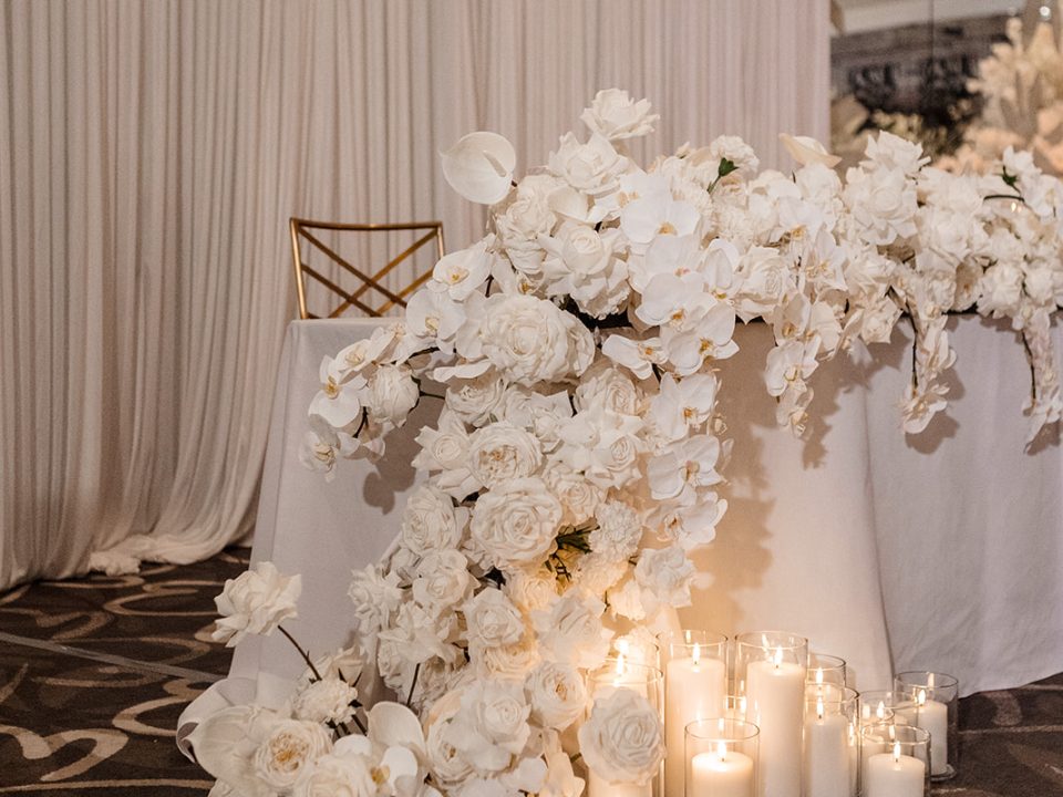 Faux orchid and rose floral table runner in all white with cylinder pillar candles at indoor reception space in Brisbane.