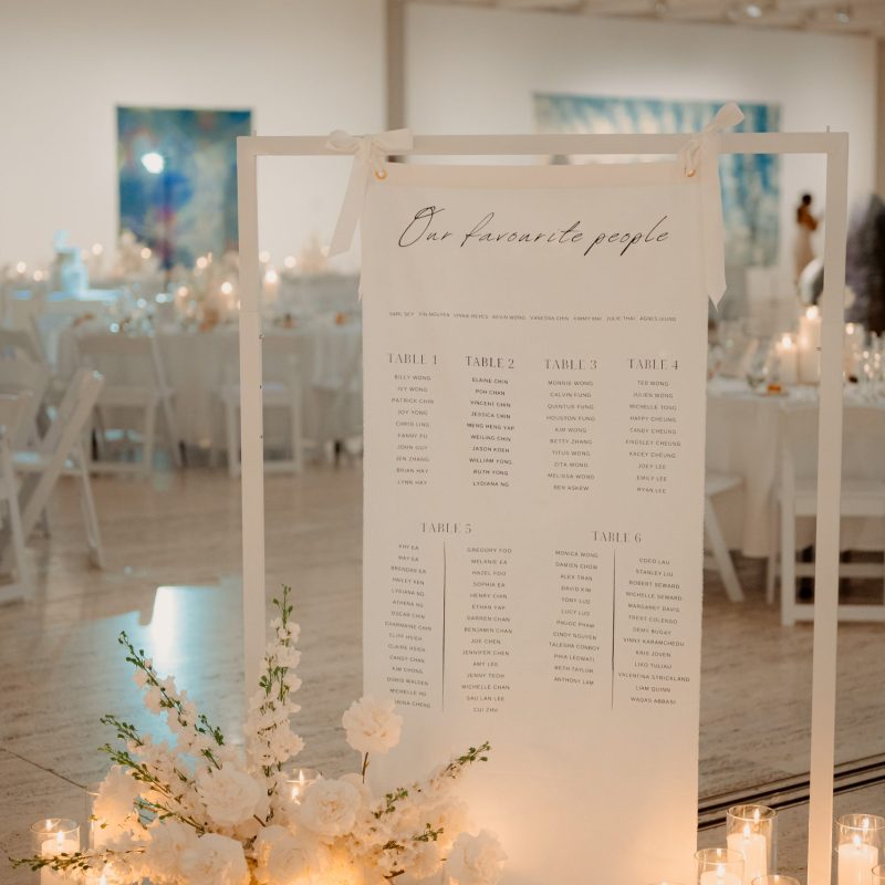 Soft linen fabric seating chart signage hung on a white display stand and styled with premium faux florals and pillar candles in glass holders.