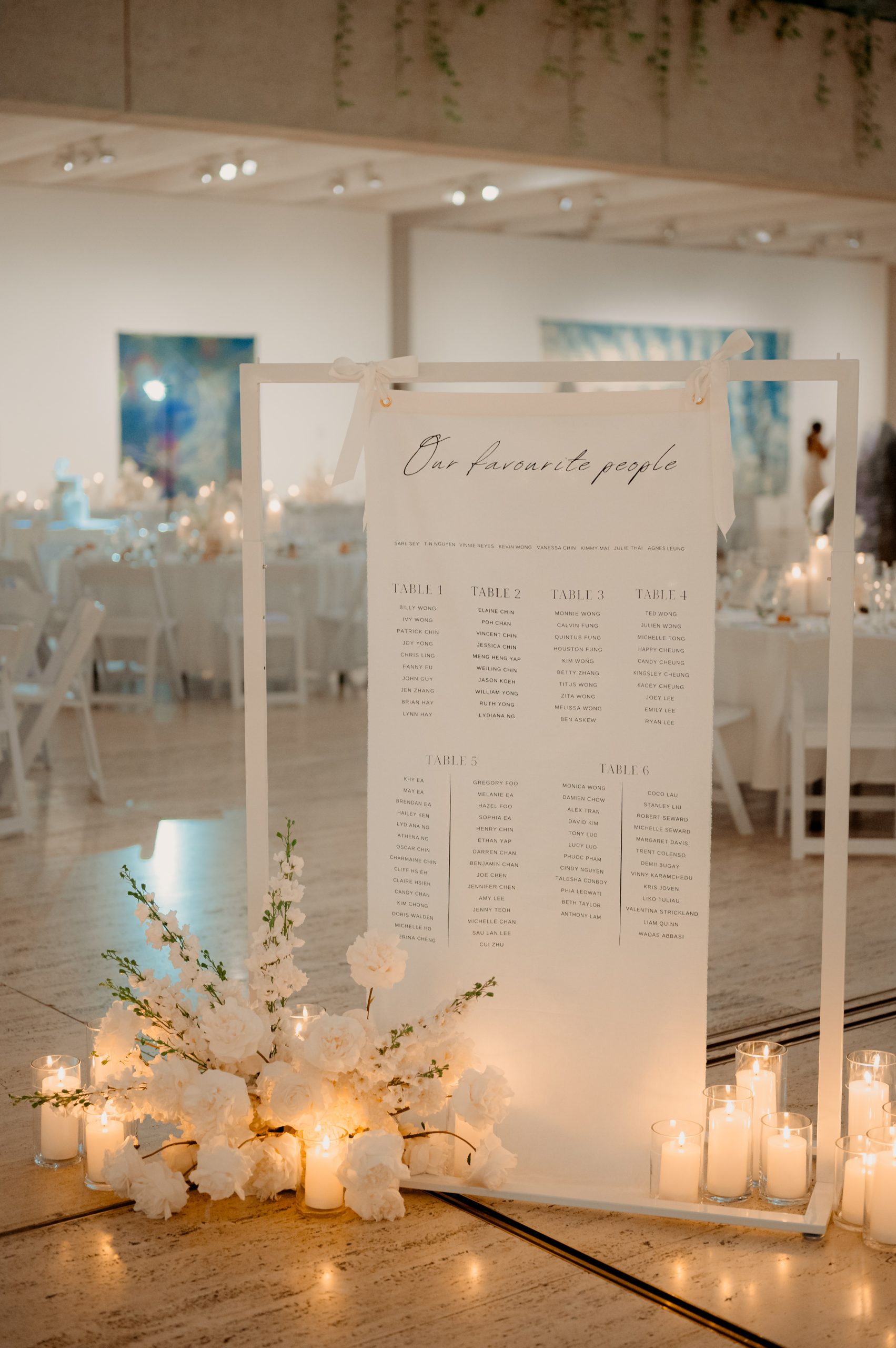 Soft linen fabric seating chart signage hung on a white display stand and styled with premium faux florals and pillar candles in glass holders.